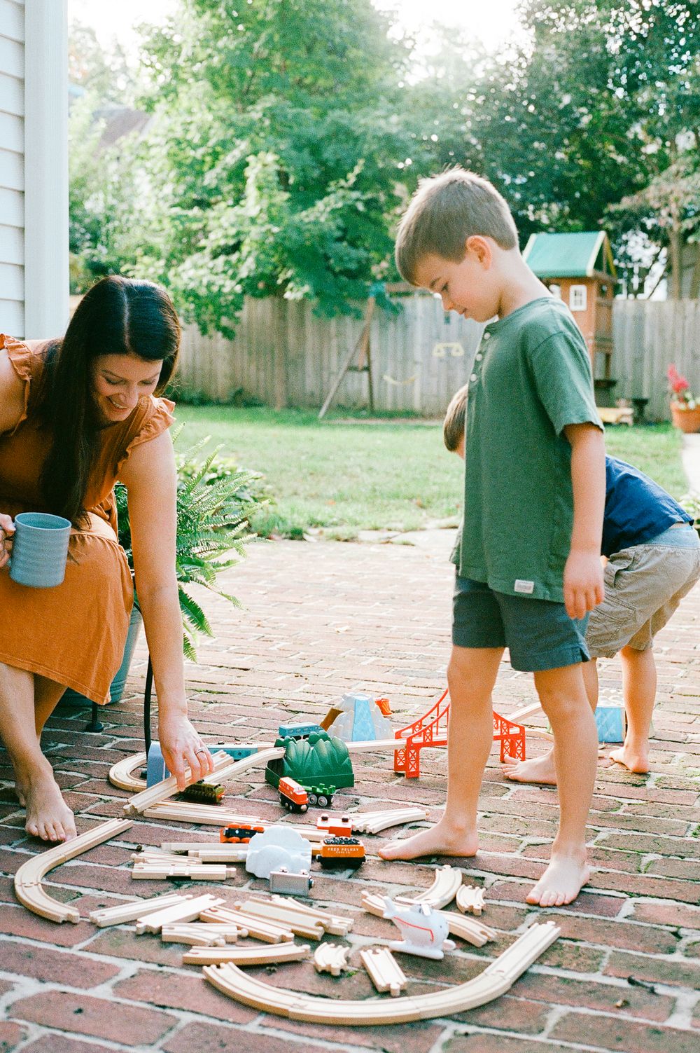 Family playing with toy train set laid out on brick patio outdoors.