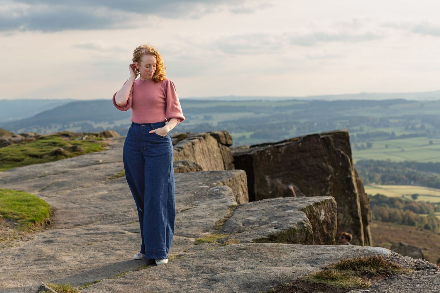 Person in pink sweater and blue pants standing on rocky cliff overlooks scenic mountain valley landscape.