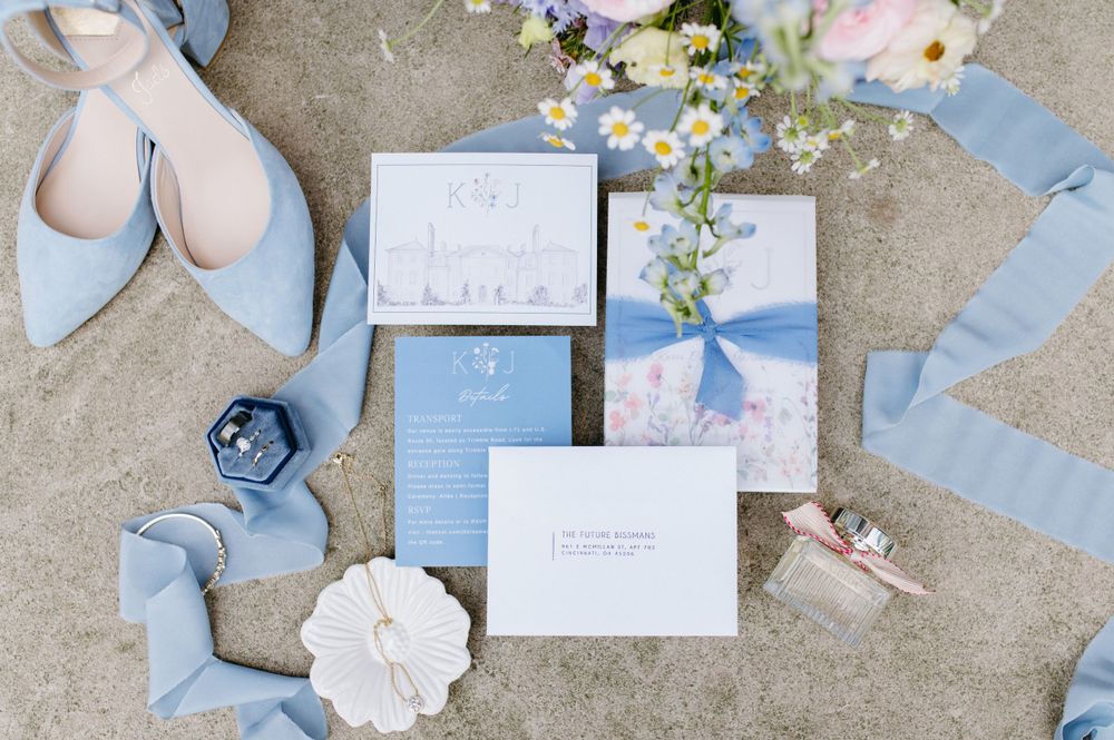 Flat lay of wedding invitations with light blue shoes and floral accents on concrete surface.