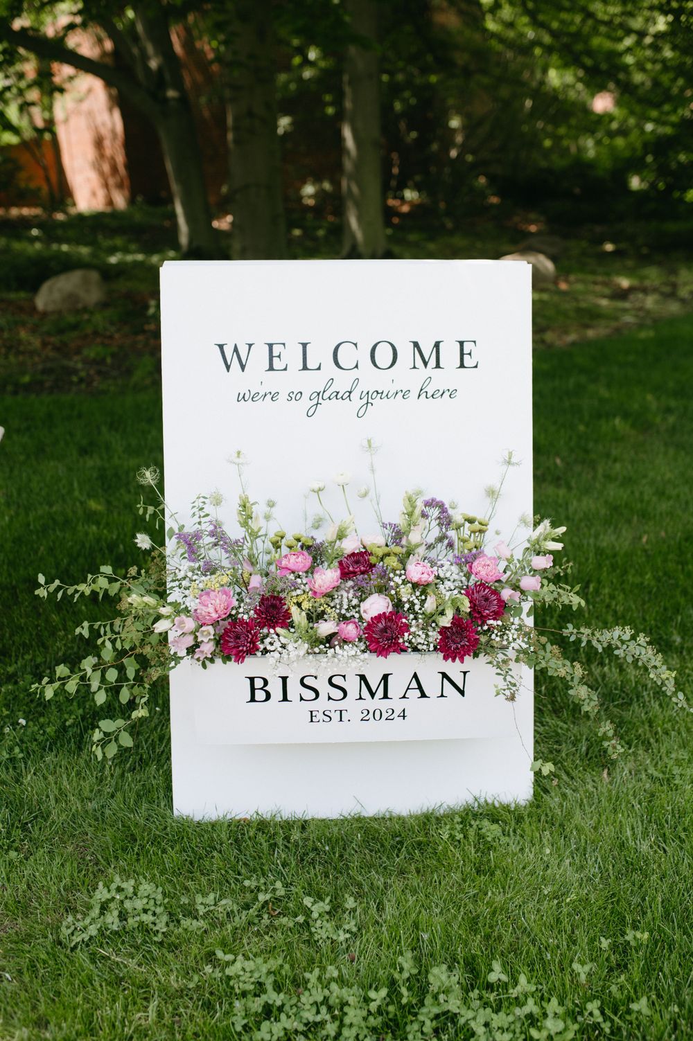 Wedding welcome sign with fresh floral arrangements and 'Bissman Est. 2024' text on white board.