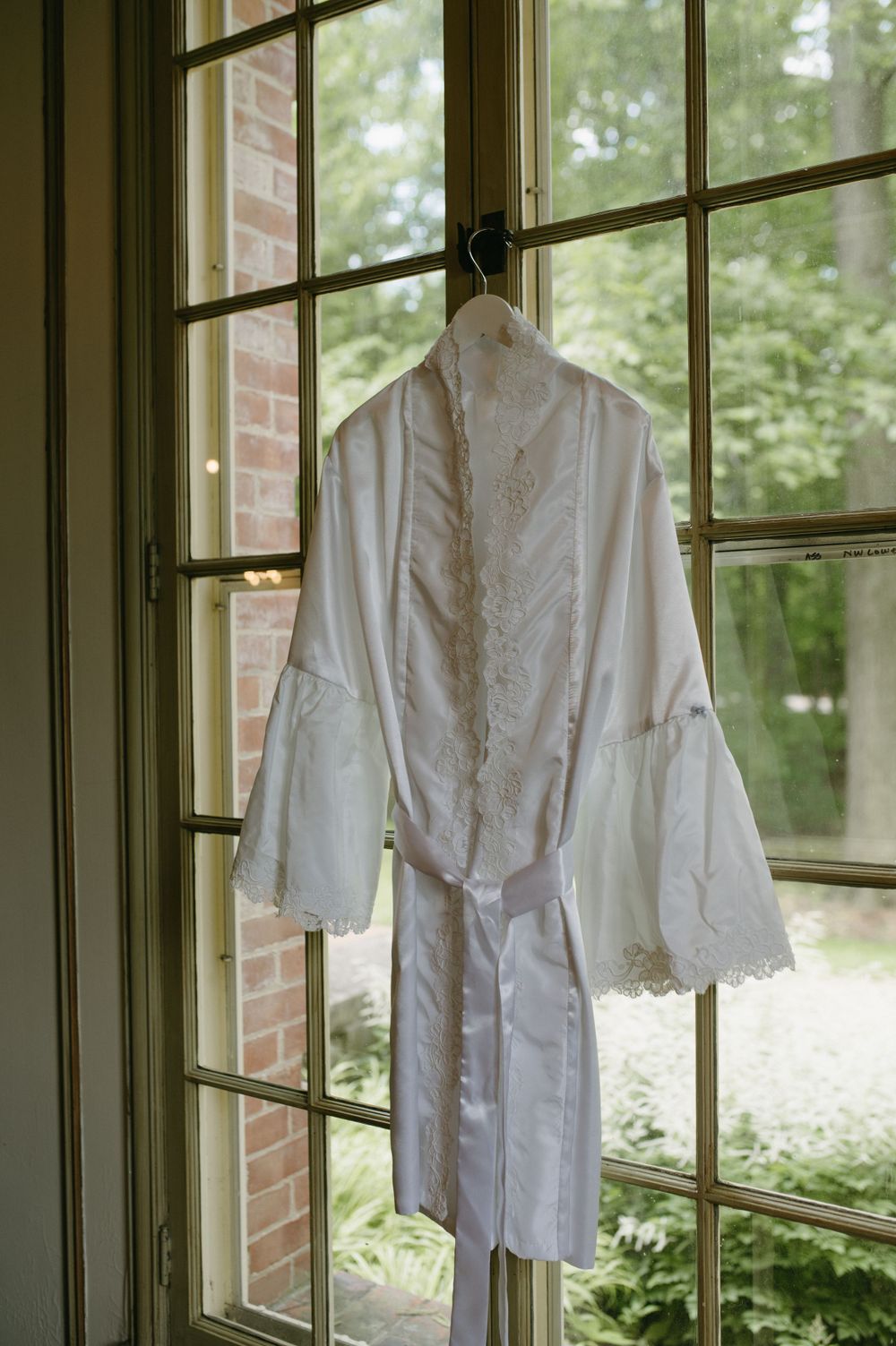 White satin bridal robe hanging in front of a window with lace trim details.