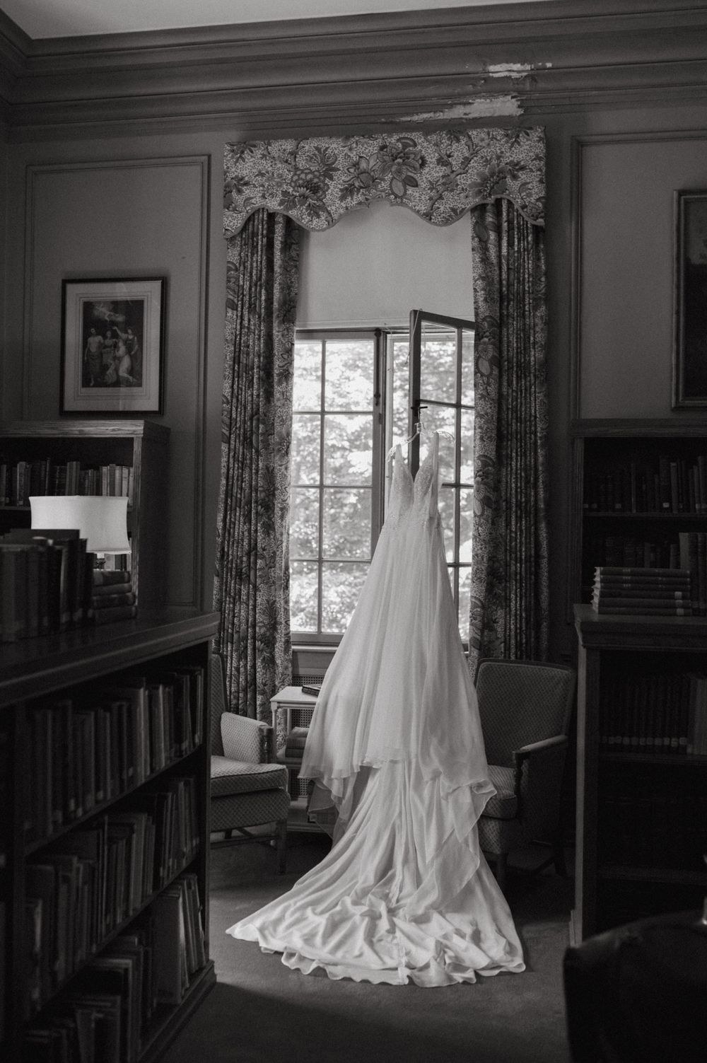 Black and white photo of an elegant wedding gown displayed in front of a window in a library setting.