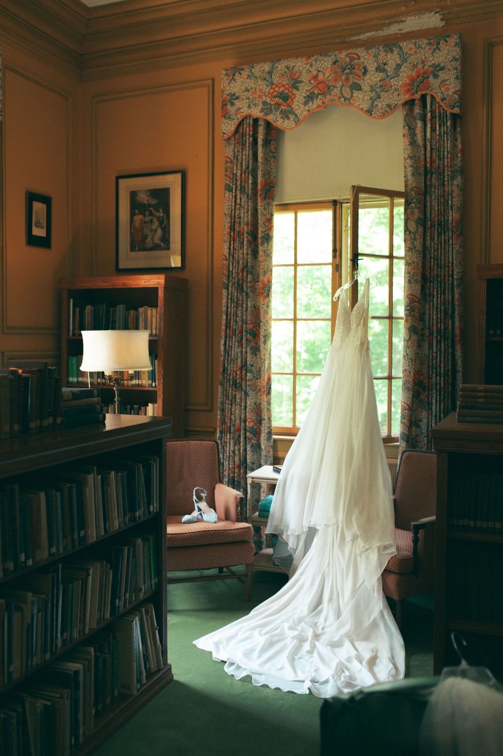 A wedding dress hanging in a library room with floor-to-ceiling bookshelves and floral curtains.