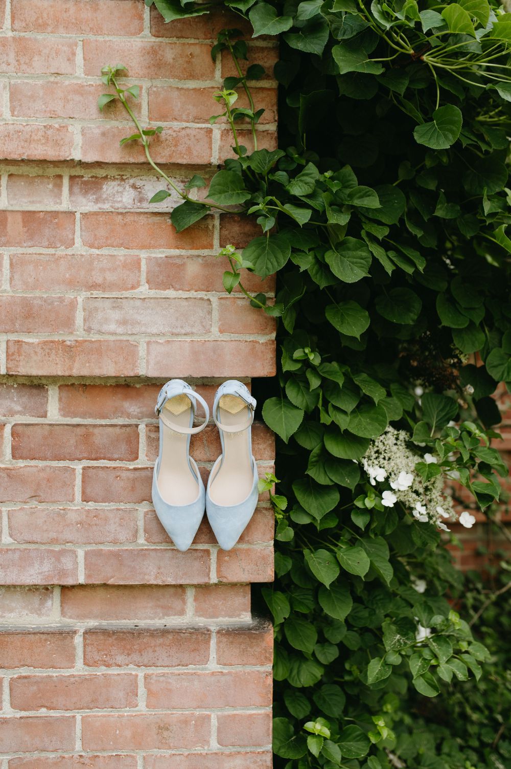 Light blue bridal shoes displayed against a rustic brick wall with climbing vines.