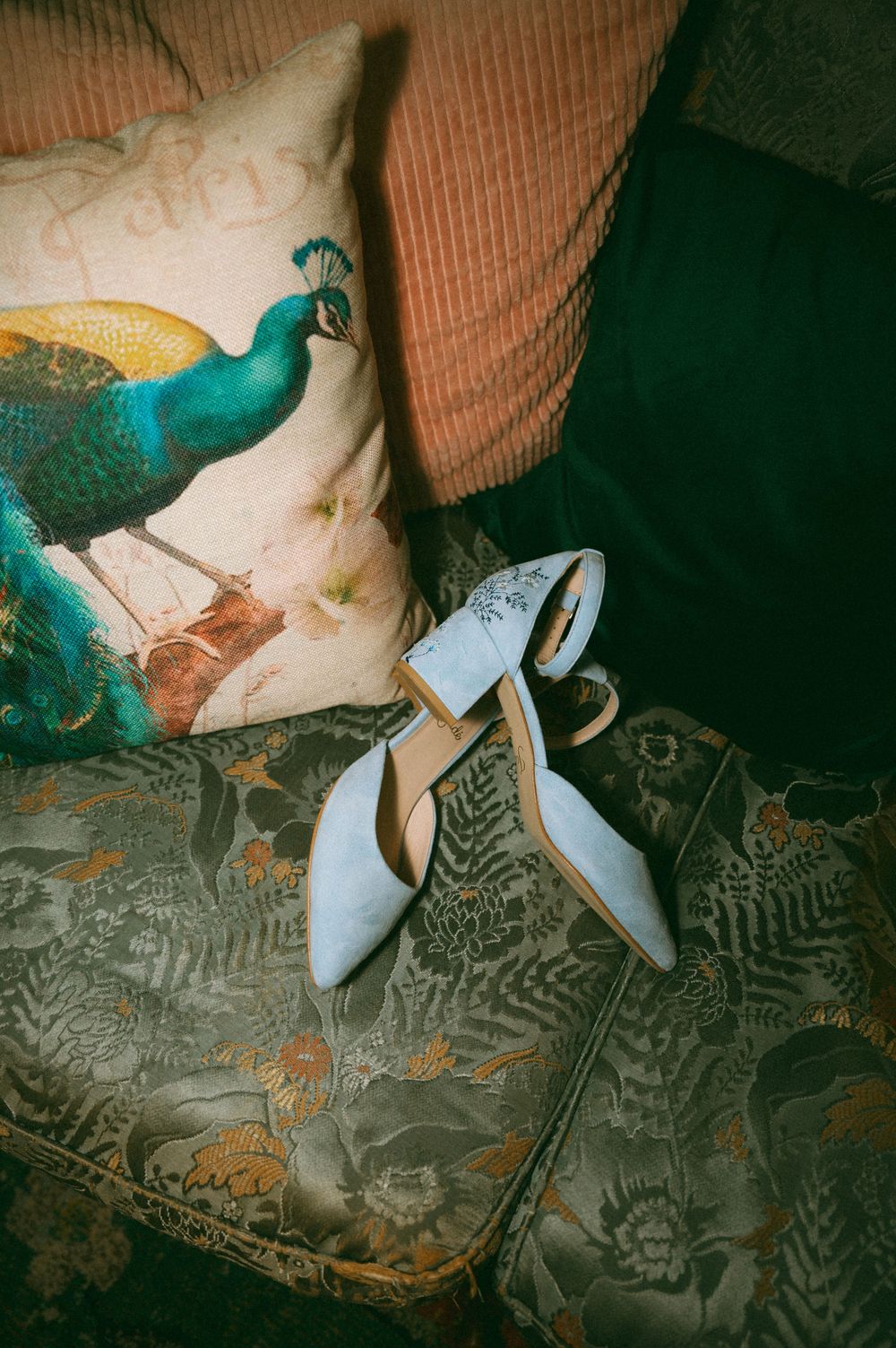 White bridal shoes next to a decorative pillow with a peacock design on a patterned couch.