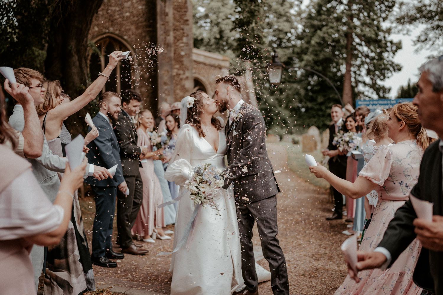 Wedding guests shower a couple with confetti along a tree-lined path during a joyful celebration.