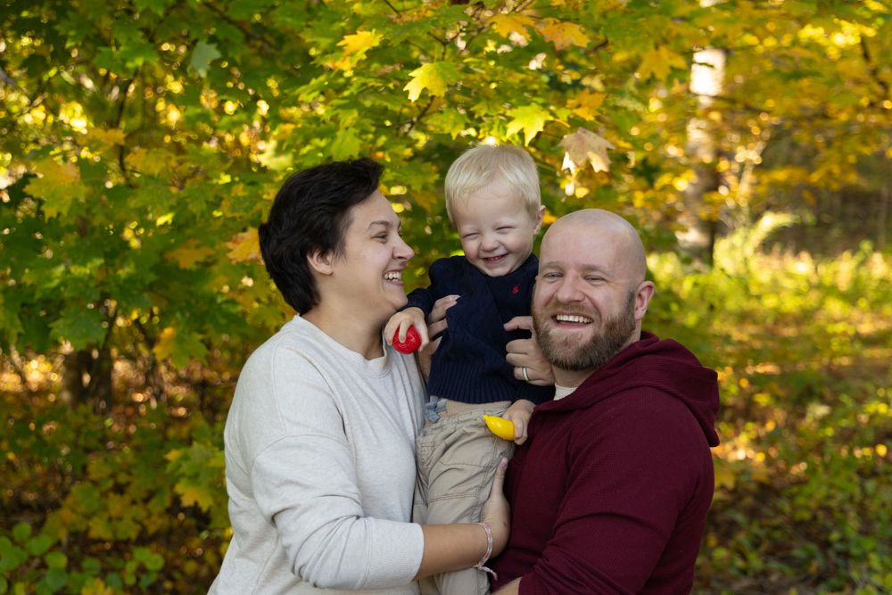 Laughing family with short-haired mother, bald father holding smiling toddler boy in autumn park setting.