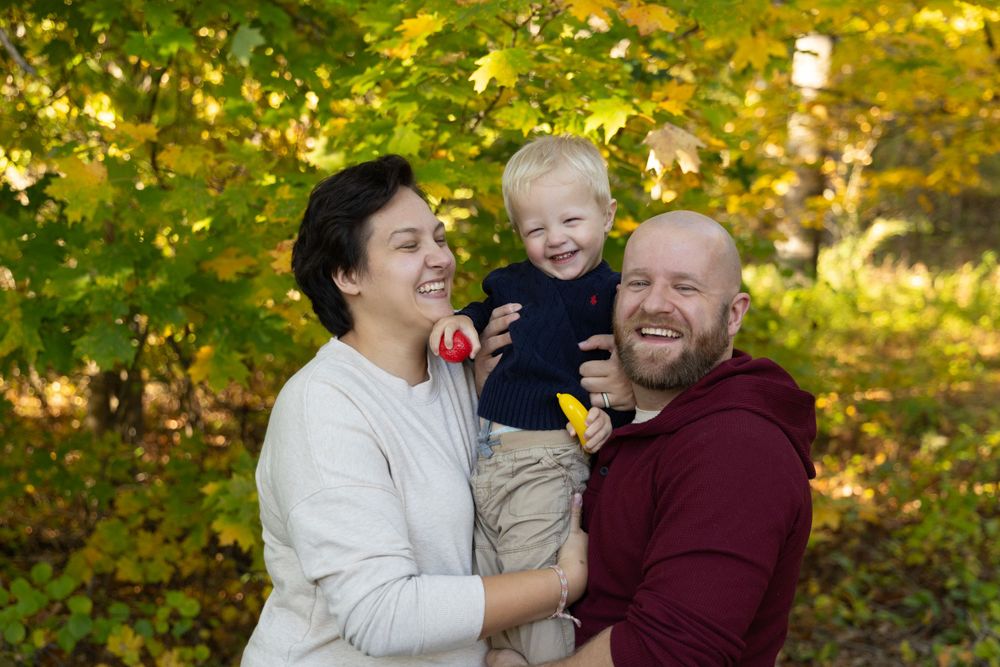 Happy family with mother, bald father, and toddler boy laughing outdoors among golden autumn foliage.