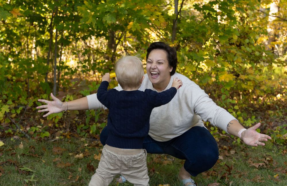 Excited mother kneeling with arms open wide as toddler runs toward her in autumn park.