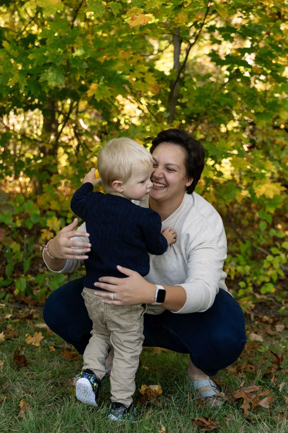 Laughing mother crouching and embracing blonde toddler boy in autumn outdoor portrait session.