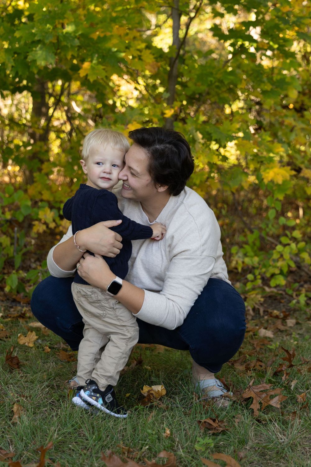Mother crouching and hugging blonde toddler boy in autumn outdoor setting.