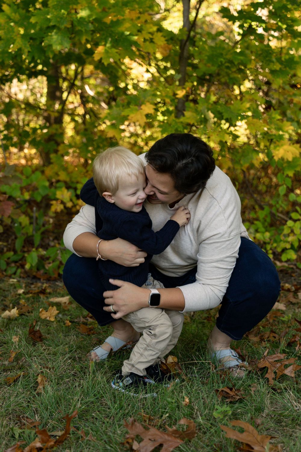 Mother kissing smiling toddler son on cheek while crouching in autumn park setting.