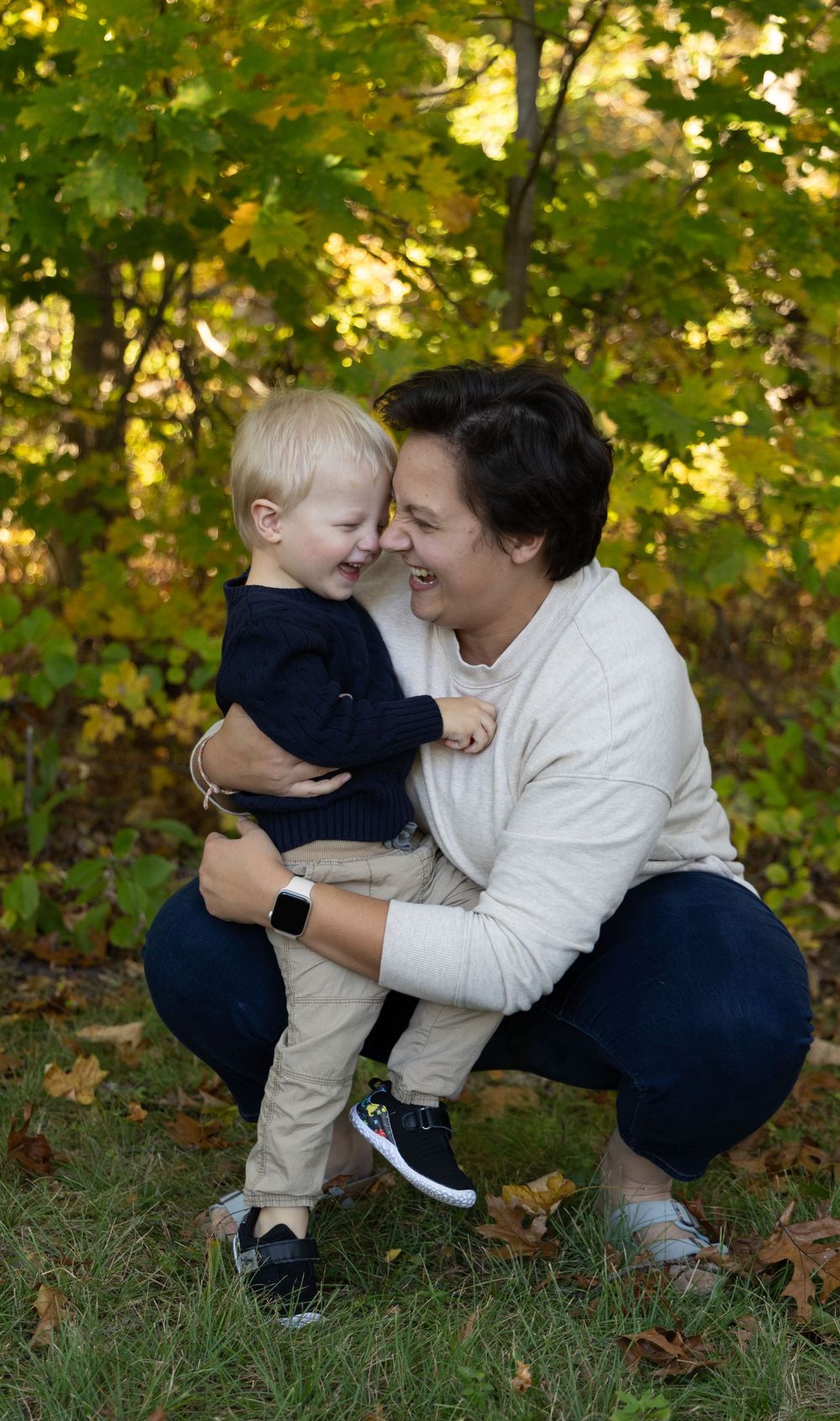 Mom holding laughing toddler boy face-to-face in fall outdoor photo session with colorful leaves.