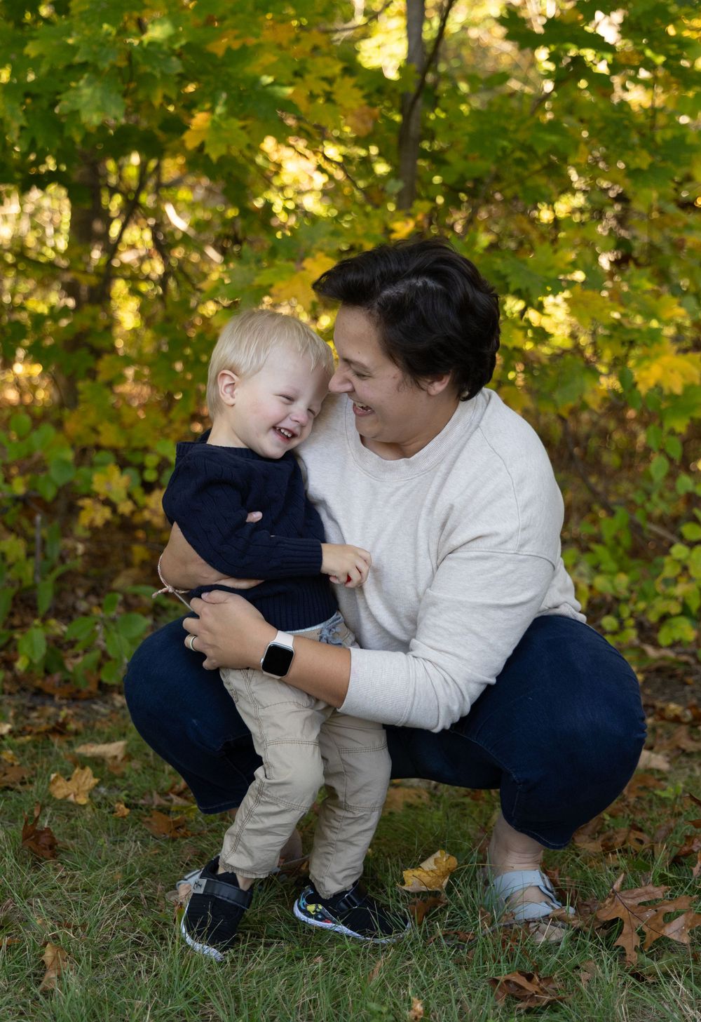 Mother crouching outdoors laughing with toddler son in autumn park with yellow foliage background.