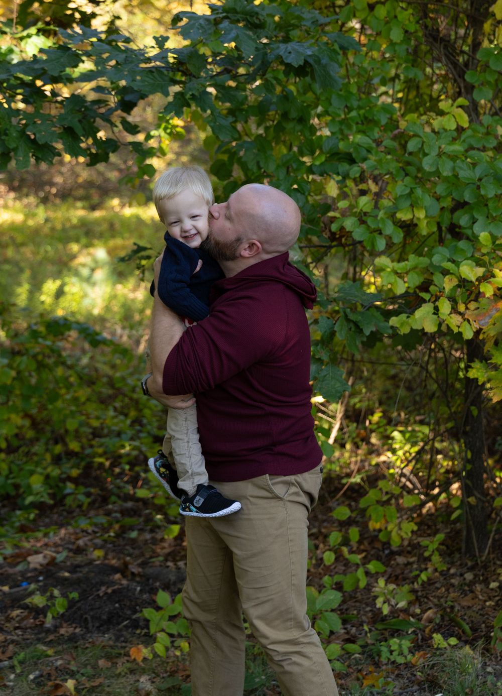 Bald father kissing and holding laughing toddler son in autumn forest.