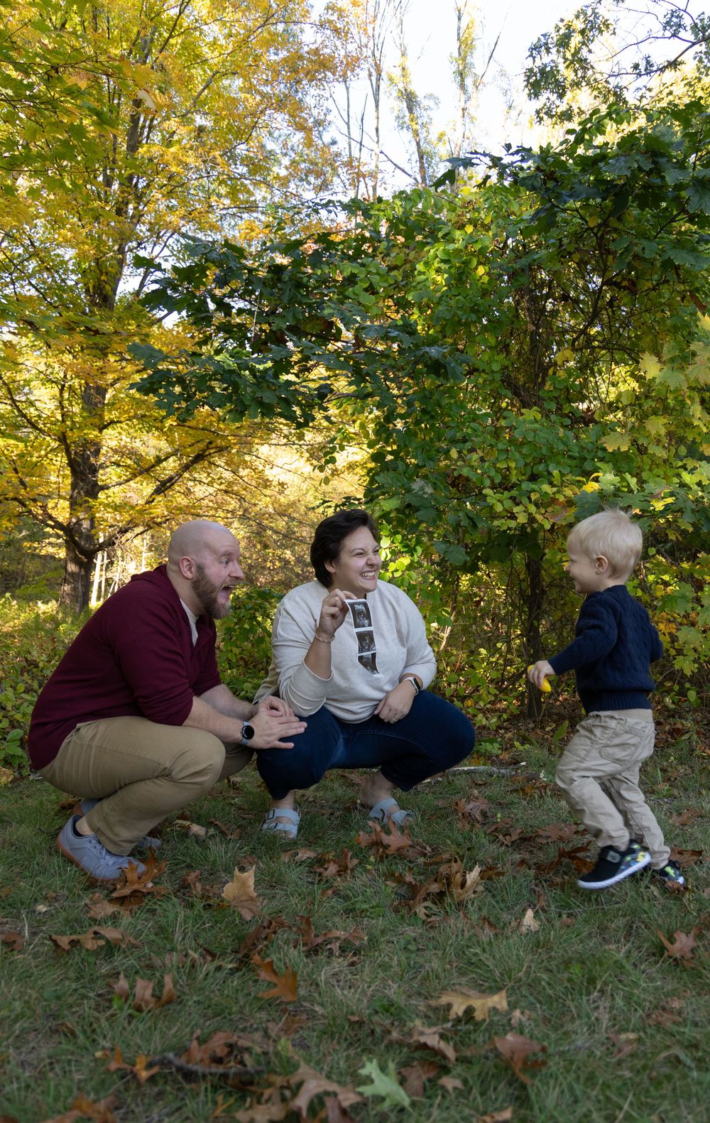 Family of three with toddler playing outdoors among colorful autumn leaves.