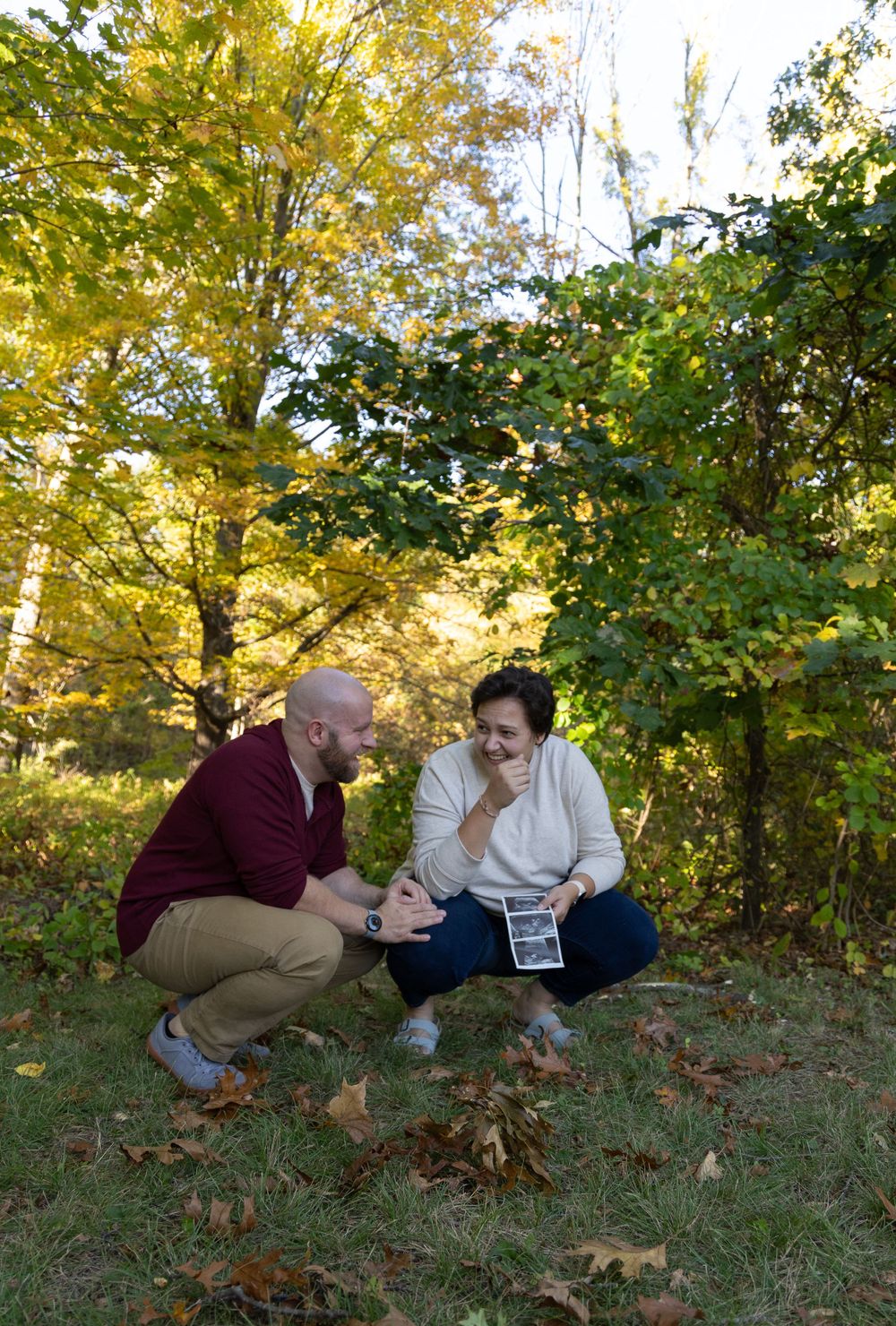 Couple crouching together laughing while holding ultrasound photo in fall park.