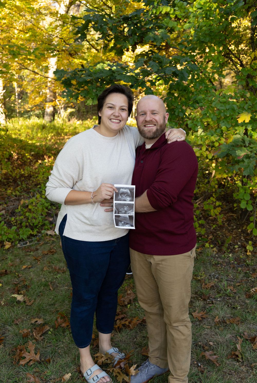 Happy couple smiling and holding ultrasound photos in fall foliage park.