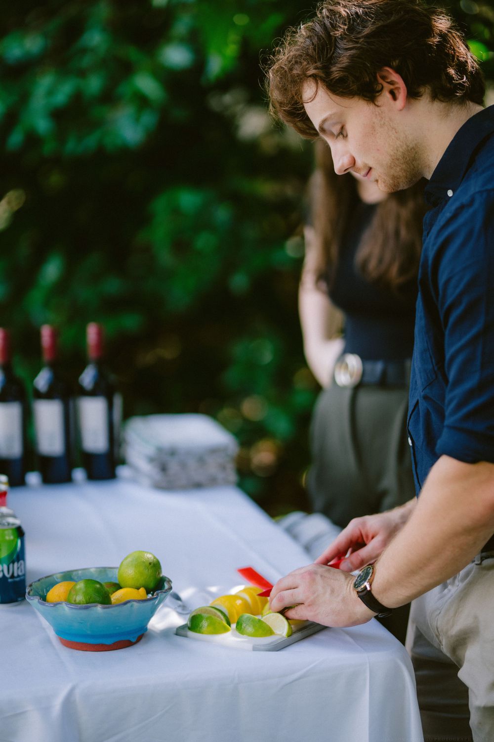 Someone preparing cocktail garnishes at an outdoor bar with wine bottles and fresh citrus.
