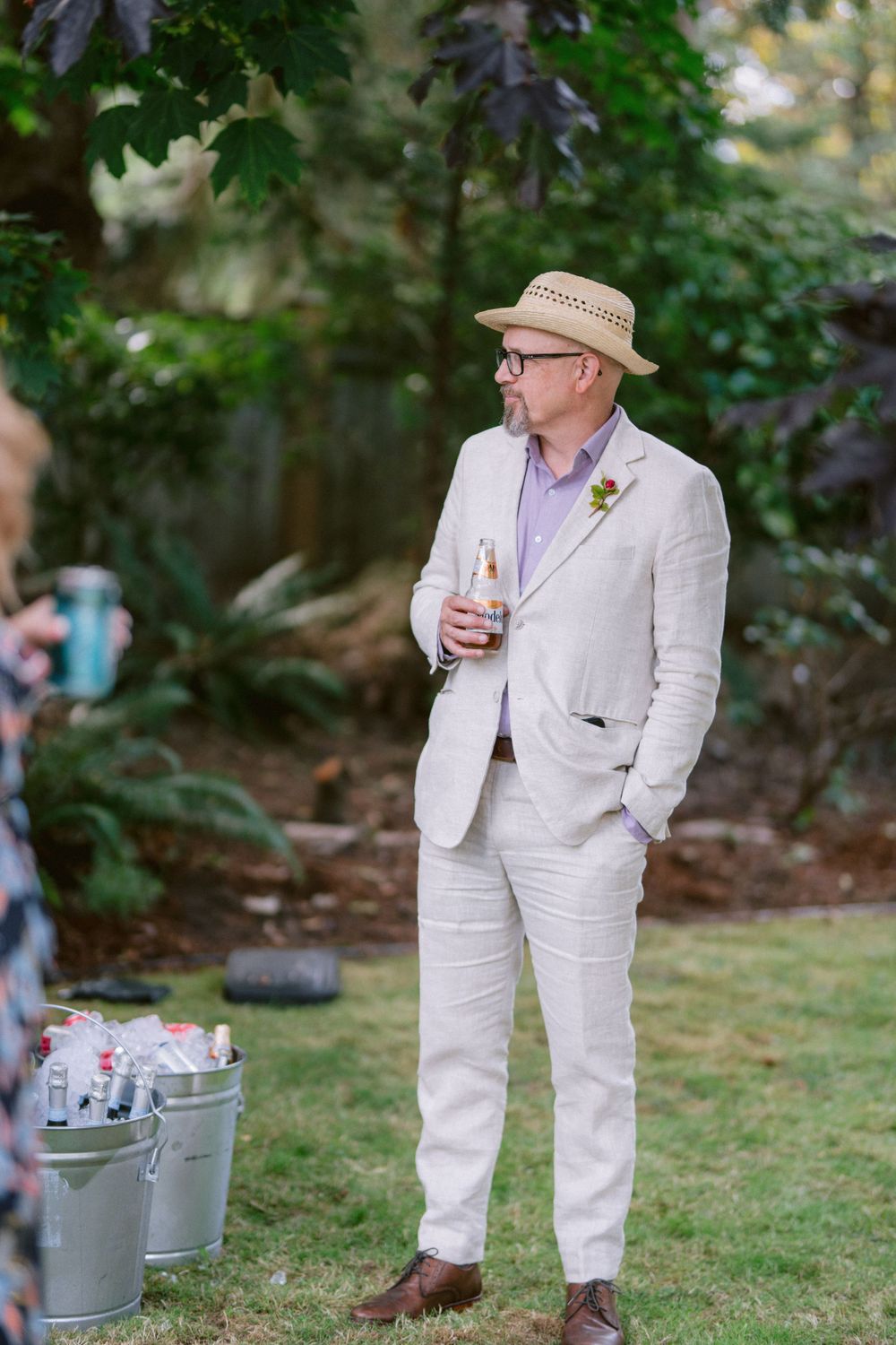 A guest in a cream-colored suit and straw hat holding a drink at an outdoor gathering.