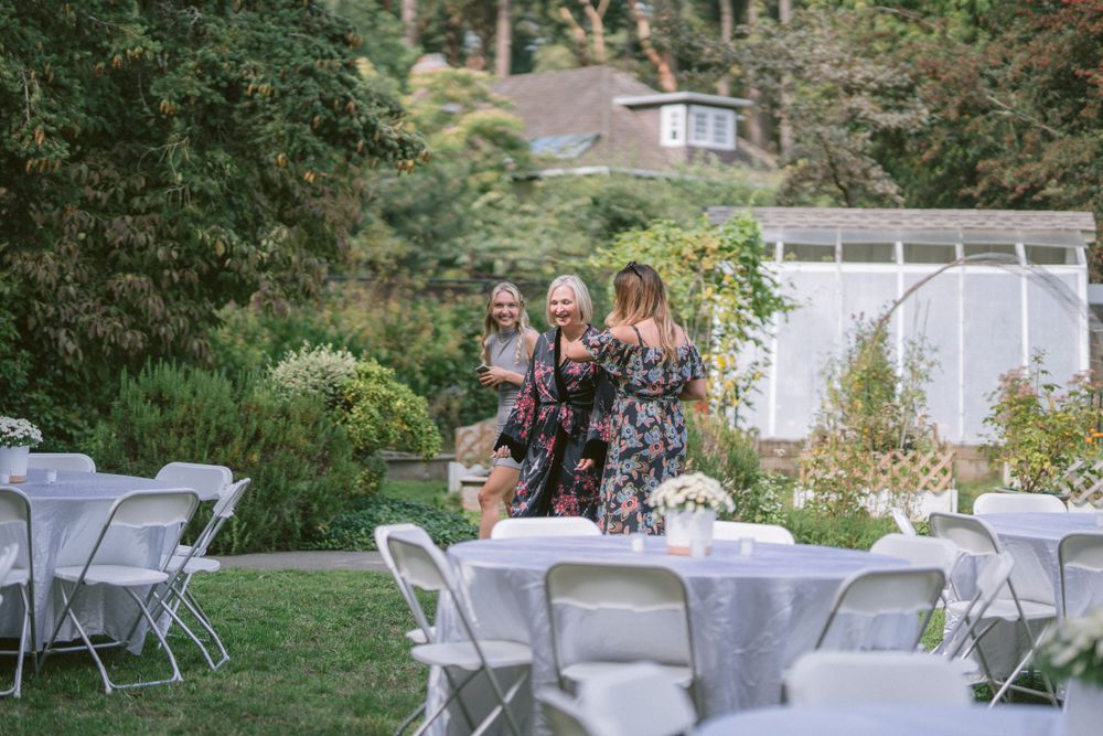 Garden party setup with white folding chairs and tables under mature trees.