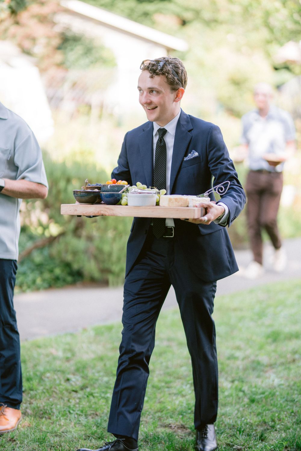 Server in navy suit carrying appetizer board at outdoor event.