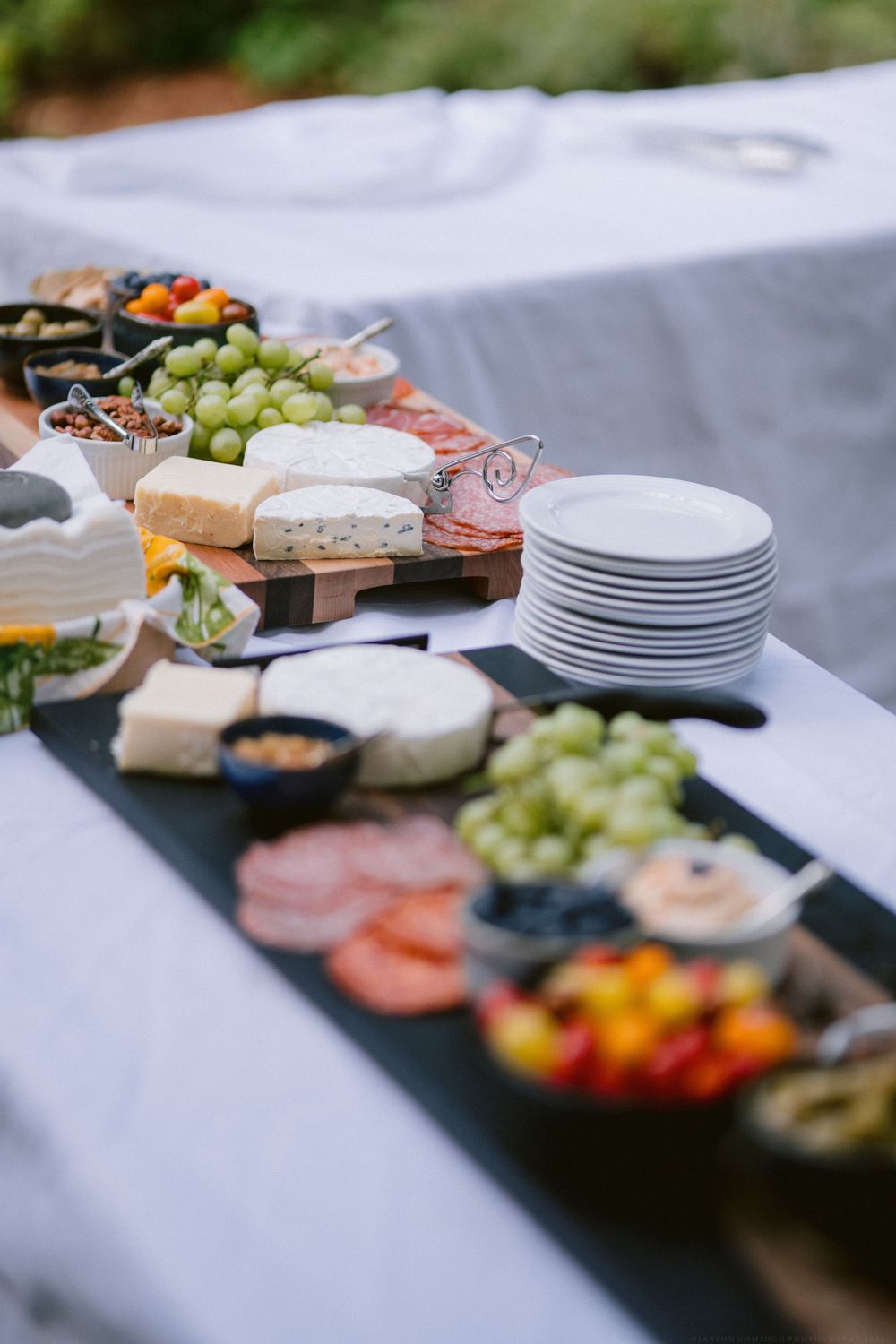Elegant appetizer spread with grapes, meats, cheeses and white plates on dark runner.