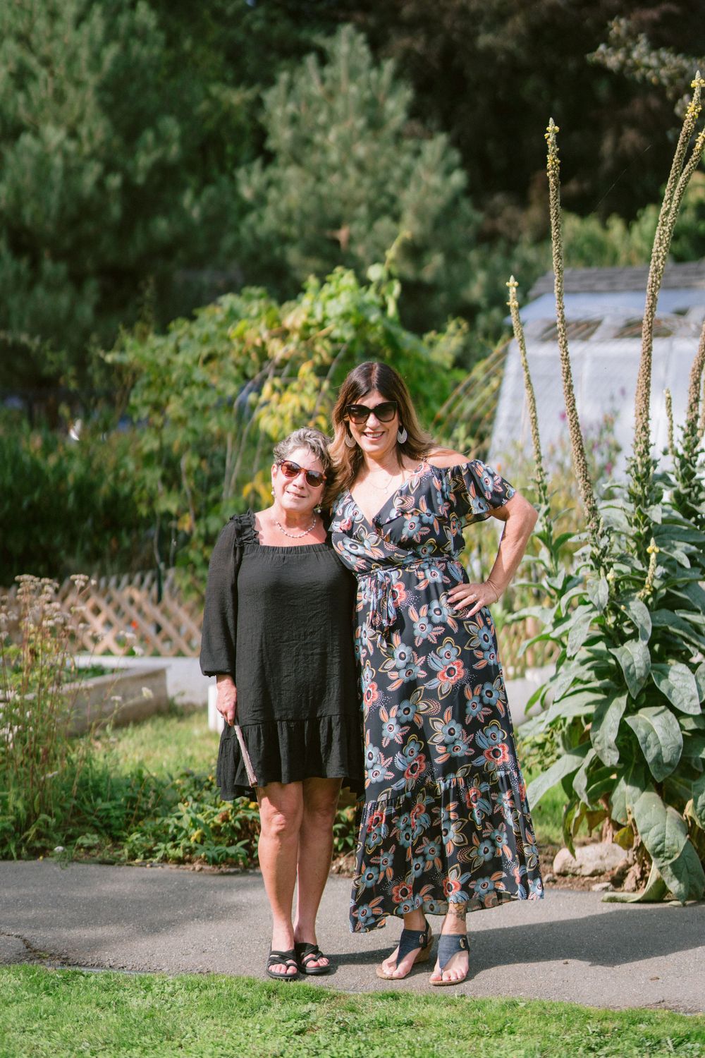 Two women stand together in a garden path, wearing contrasting summer dresses with a maxi floral dress and black dress.
