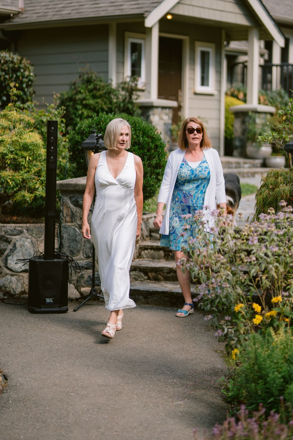 Two women walking along a garden path in summer dresses with a craftsman style house in background.