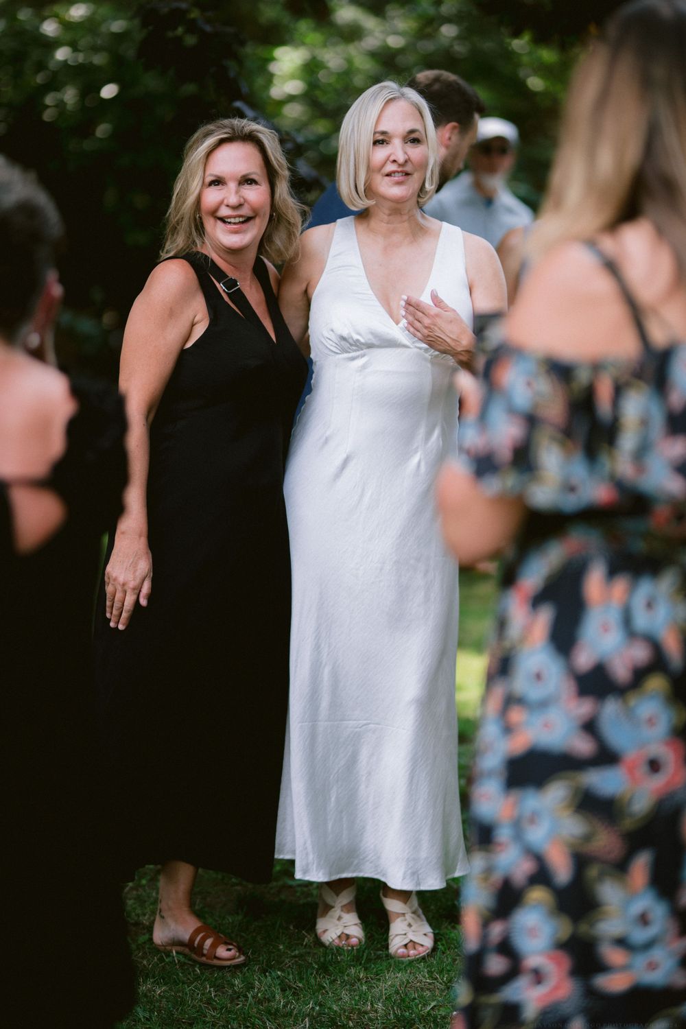 Friends pose together at outdoor summer gathering, one wearing white dress and other in black.