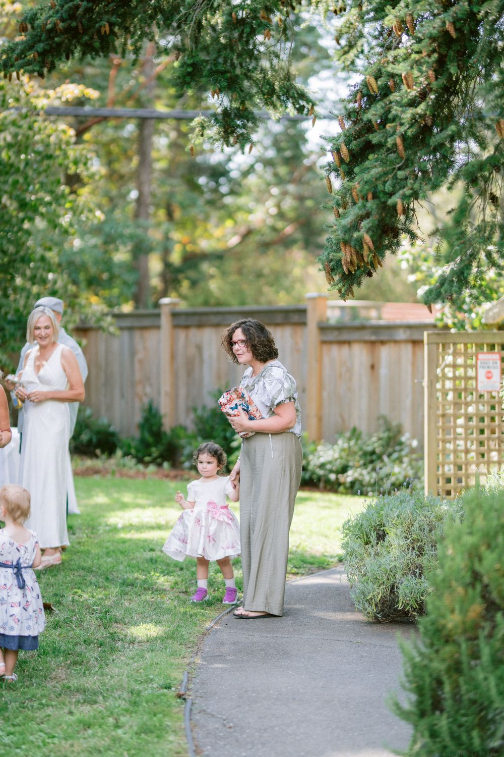 Garden party attendees gather on a paved pathway surrounded by lush greenery and wooden fencing.