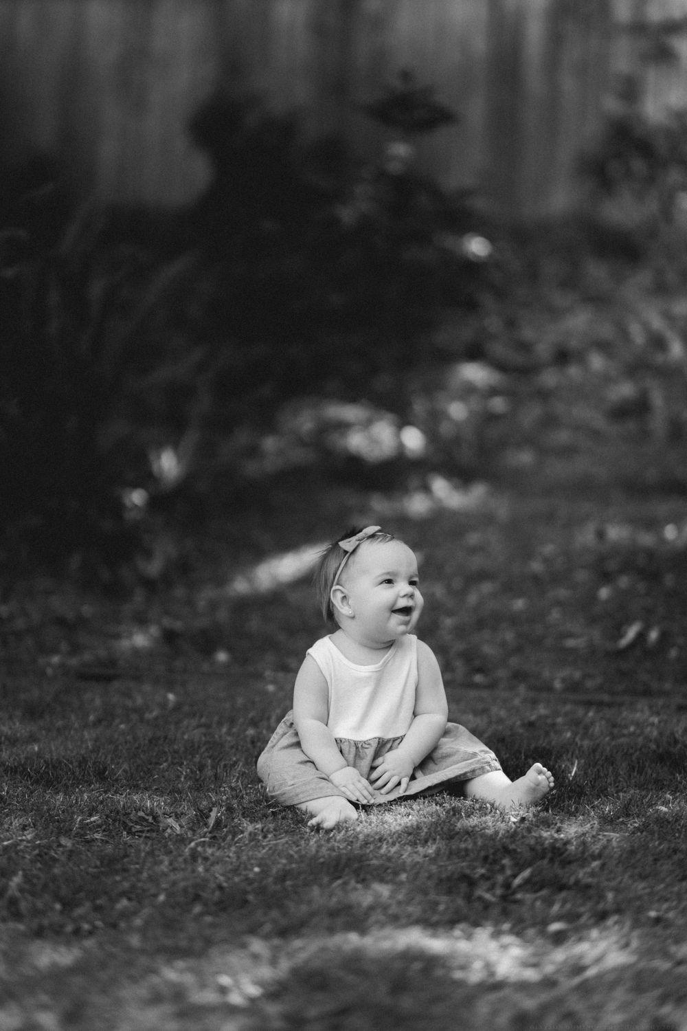 Black and white photo of a small child sitting and laughing on grass in a backyard setting.