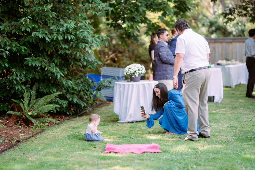 Casual outdoor gathering with people mingling around tables and chairs on a grass lawn.
