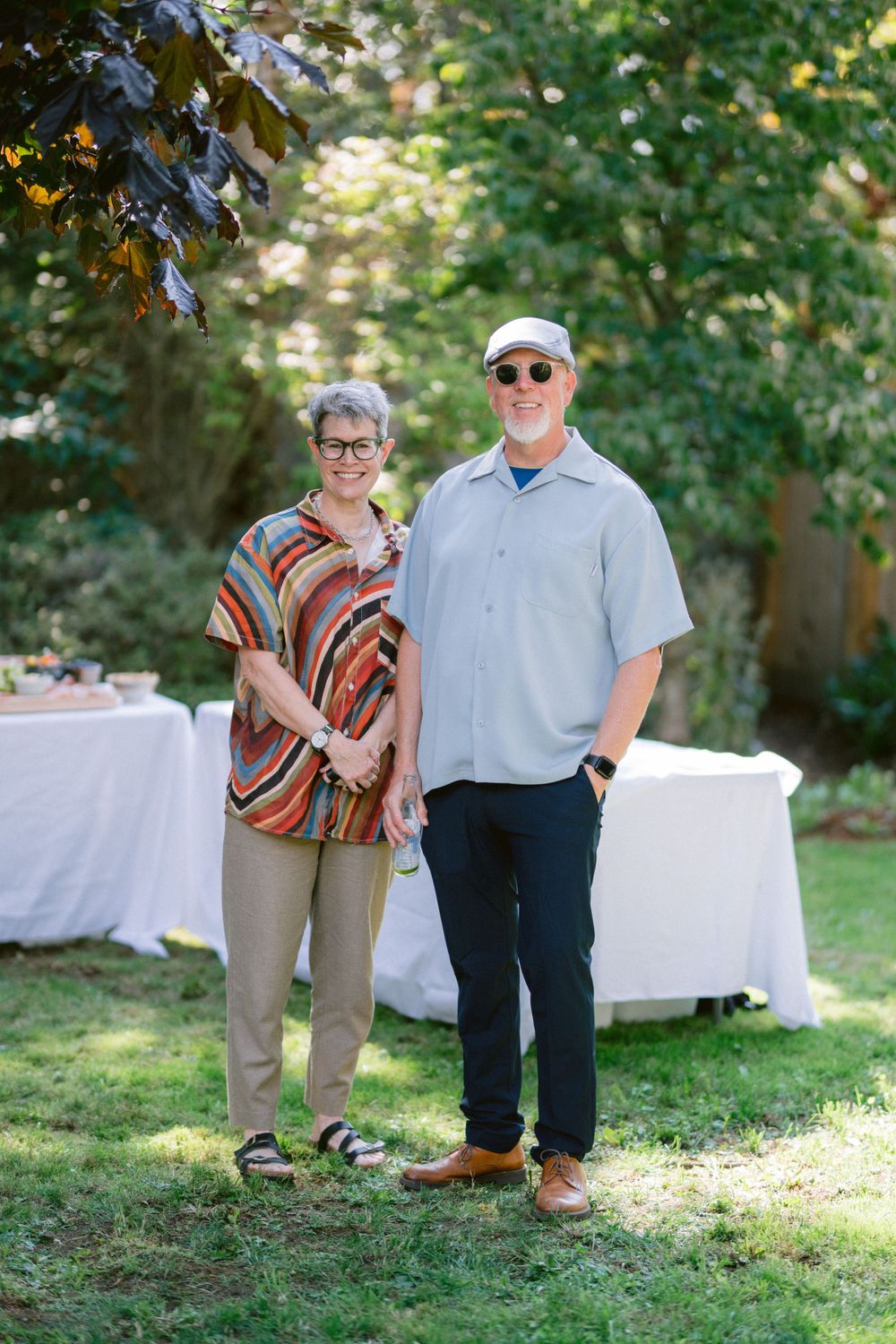 Couple poses together in casual attire near white-clothed tables at an outdoor event.