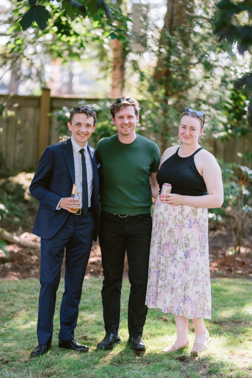 Three friends pose together at an outdoor gathering holding drinks in a leafy backyard setting.
