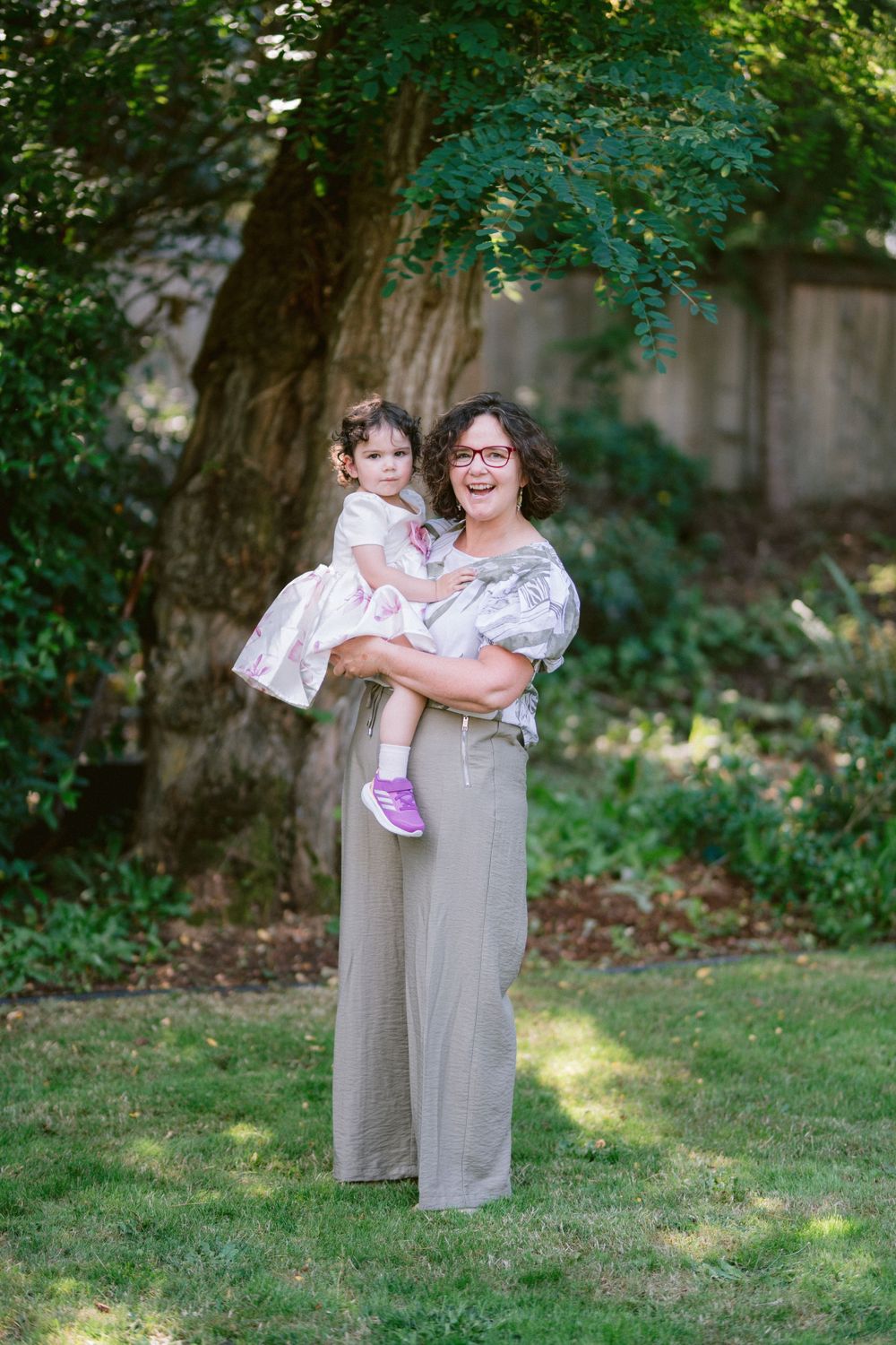 A parent holds a small child wearing a pink dress in a shaded garden setting.