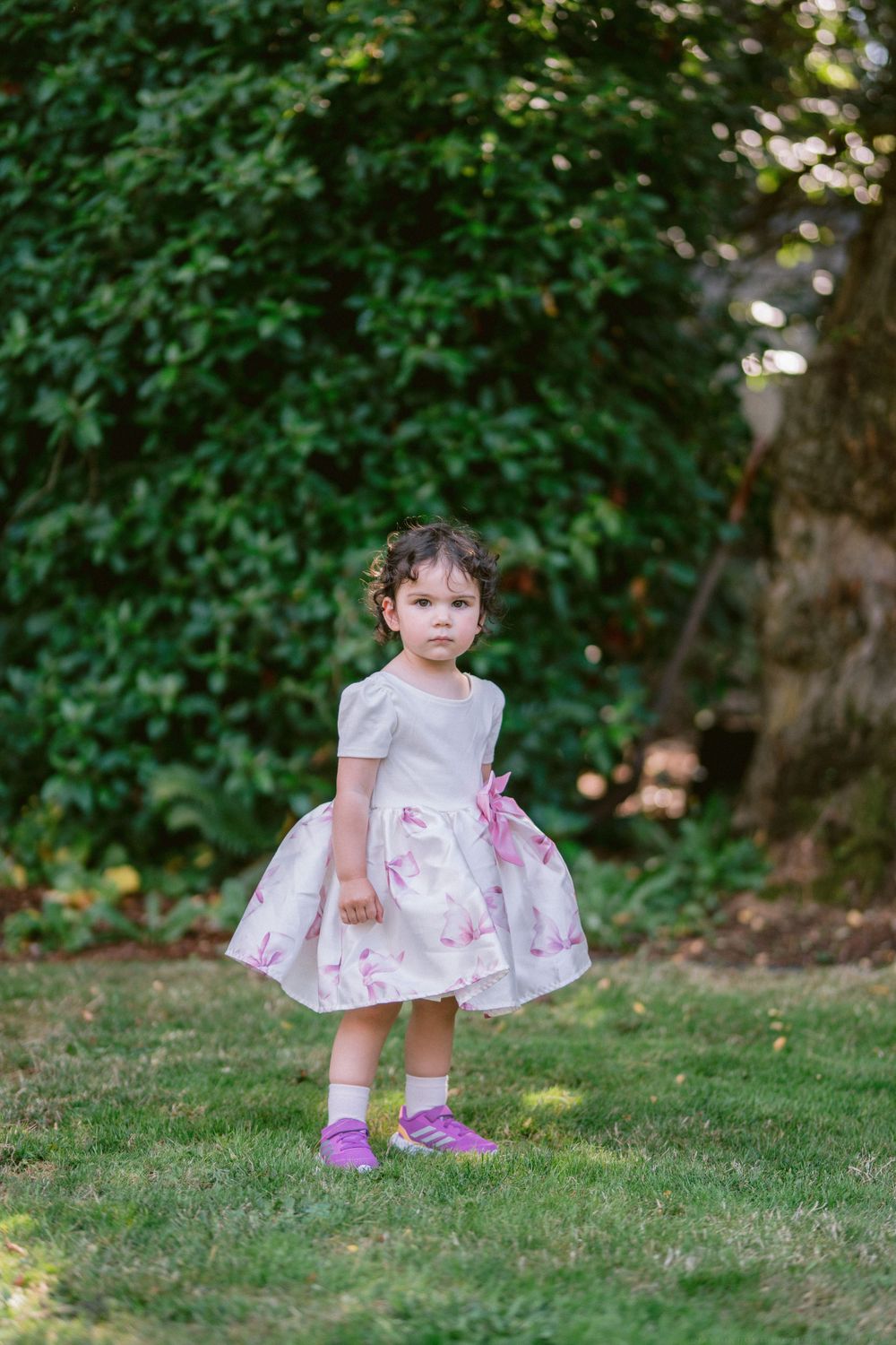 A small child in a floral dress and purple shoes stands on grass with trees in the background.