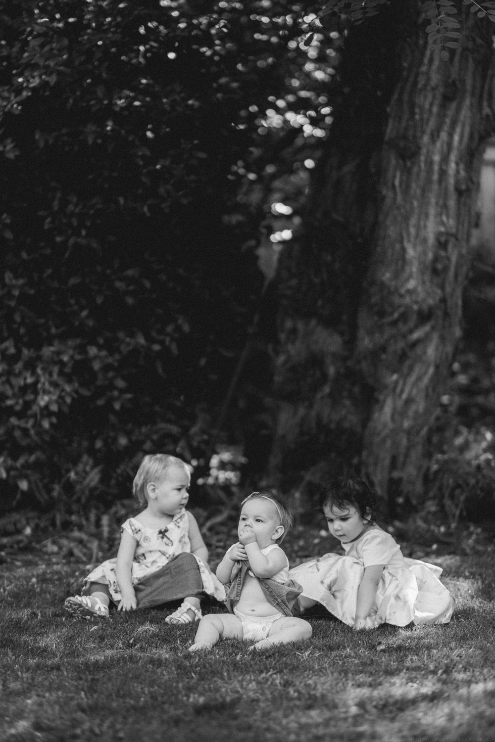 Three young children sit together on grass in a black and white photo under trees.