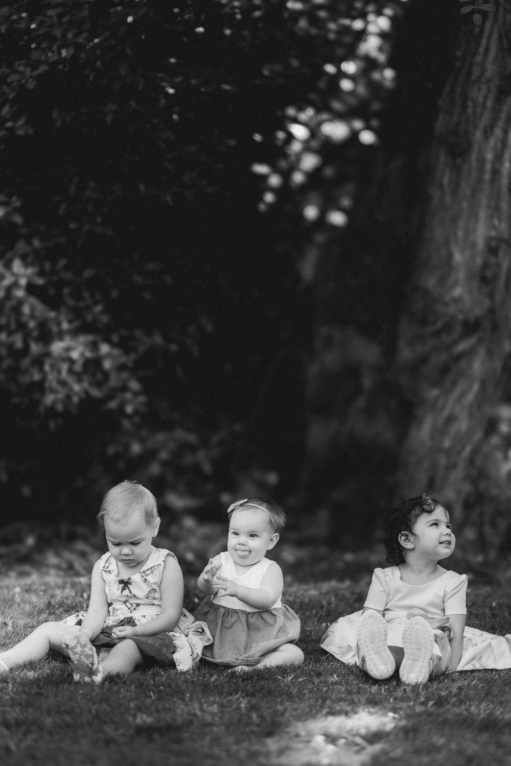 Three babies sit together on grass in black and white photography.