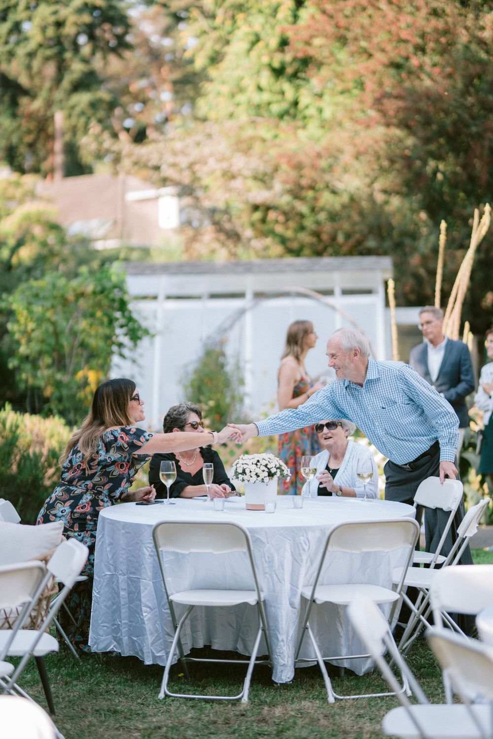 People interact around tables at an outdoor garden party with white tablecloths.