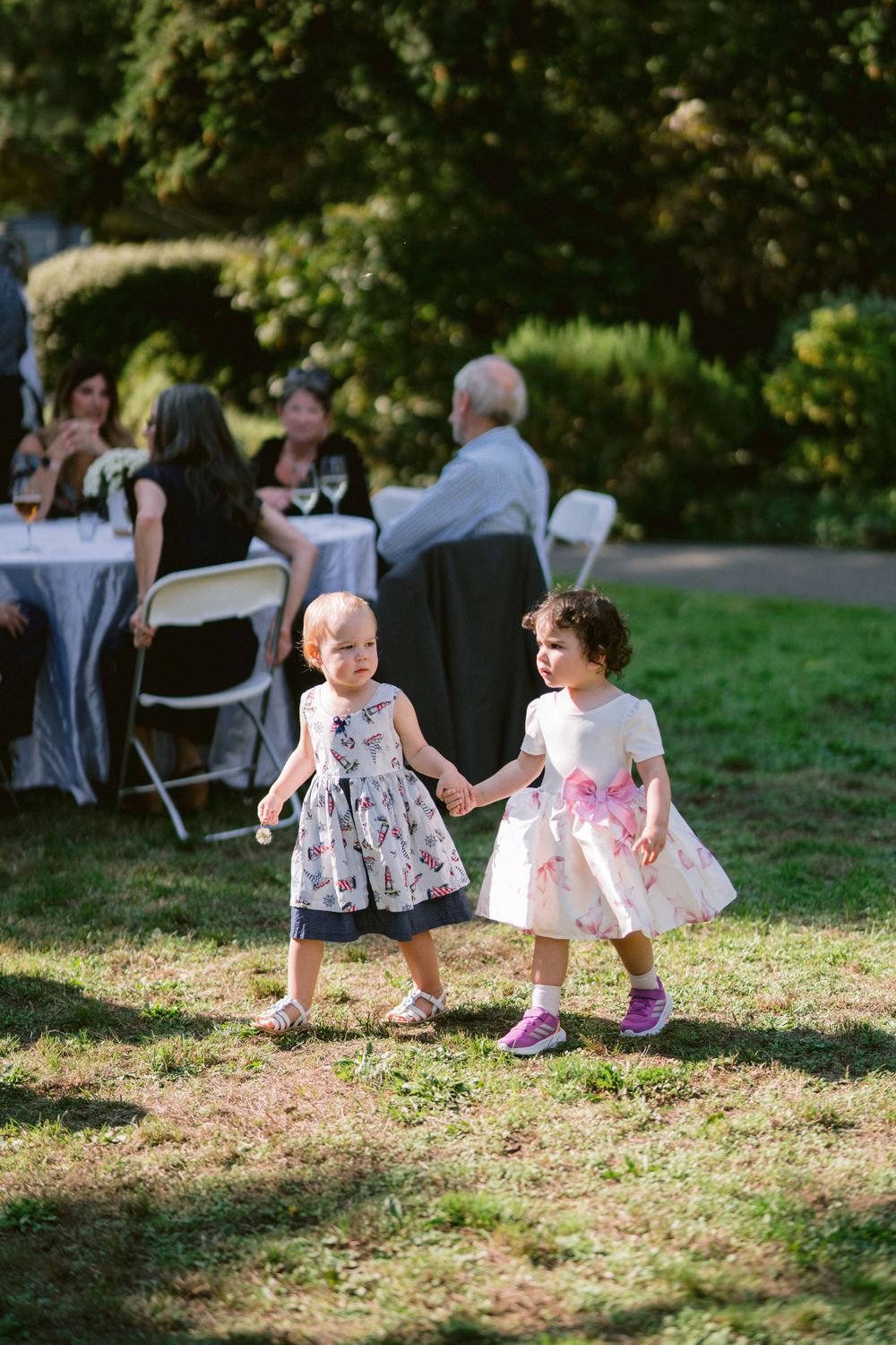 Two children in floral dresses dance and play together at an outdoor garden party reception.