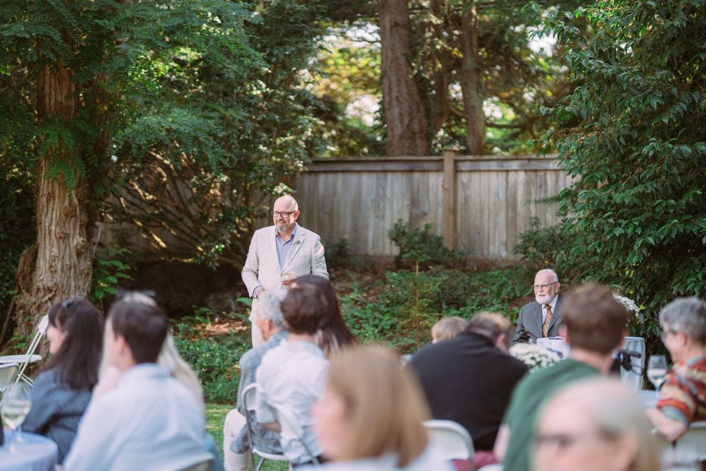 Guests seated at outdoor reception listen attentively to speech during wedding celebration.