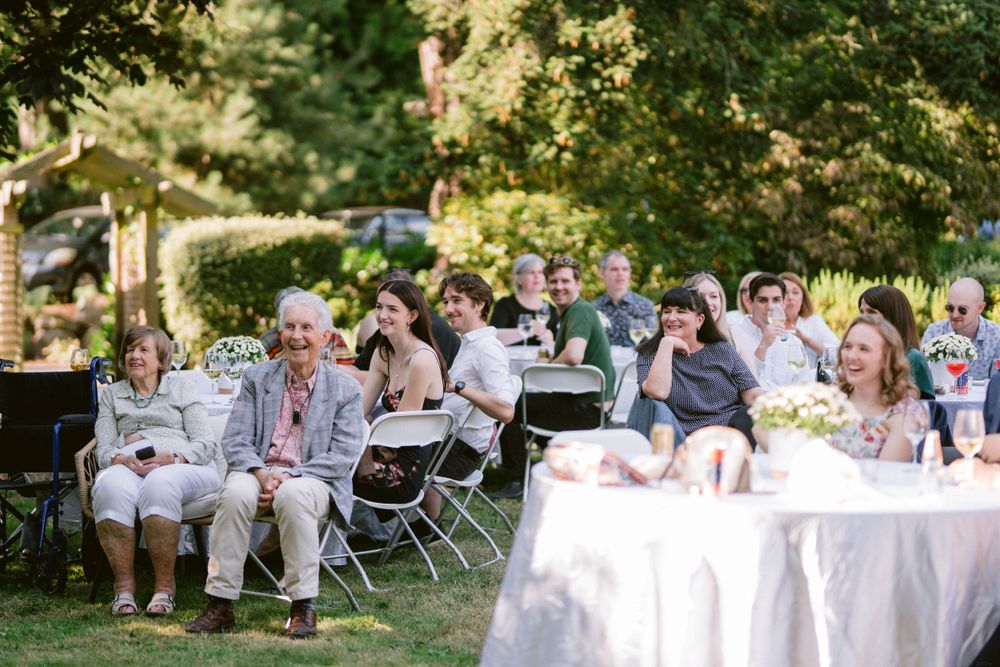 Guests seated at outdoor wedding reception with white tablecloths and folding chairs.
