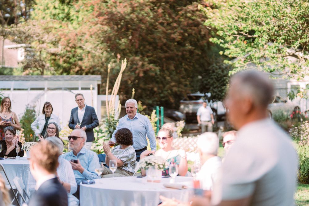 Guests enjoy an outdoor garden reception at round tables with white tablecloths in a sunlit backyard setting.