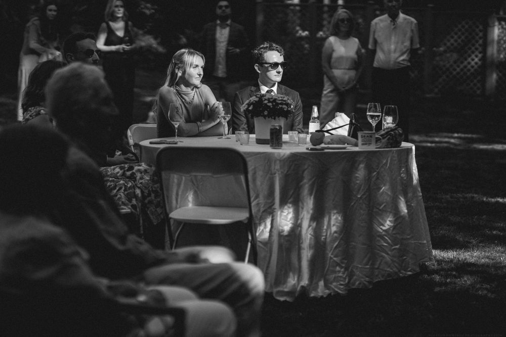 Black and white image of guests seated at a decorated table during an outdoor event.