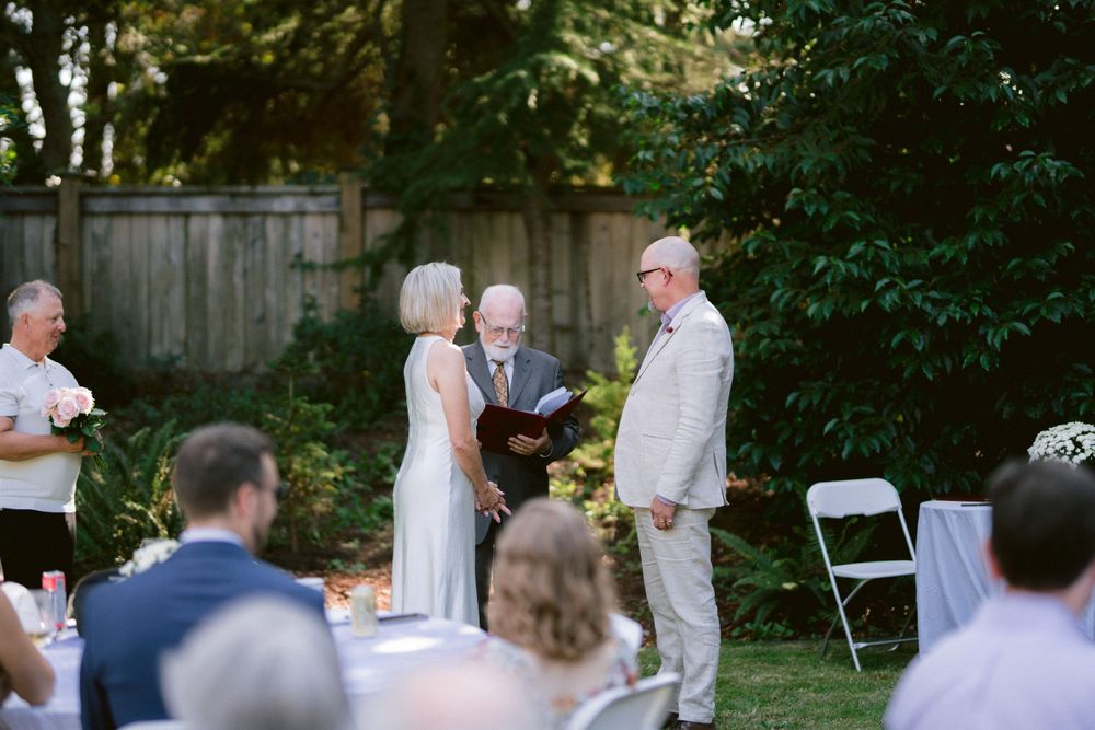 Outdoor garden wedding ceremony with guests seated on white chairs.