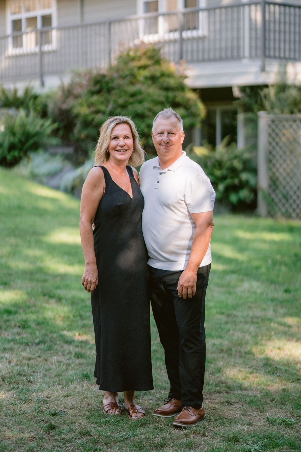 A couple poses together on a landscaped lawn in front of a house with a balcony.