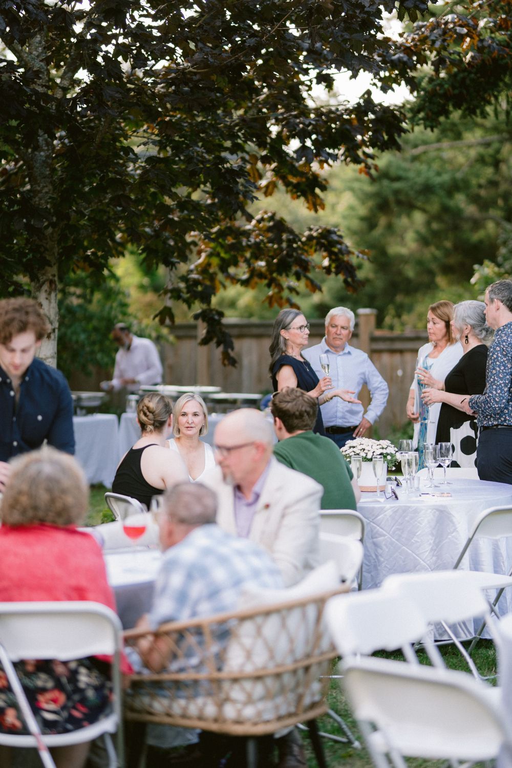 Garden party guests seated at round tables covered in white linens under tree canopy.