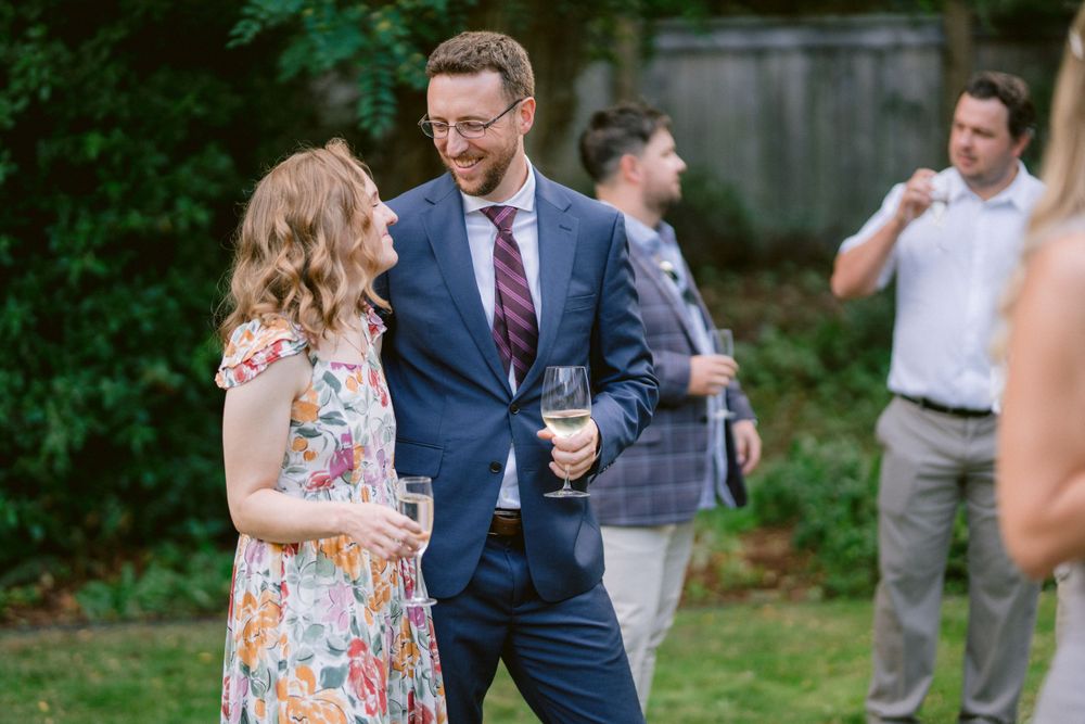 Two guests mingle at an outdoor garden party, with one wearing a floral dress and another in a navy suit.