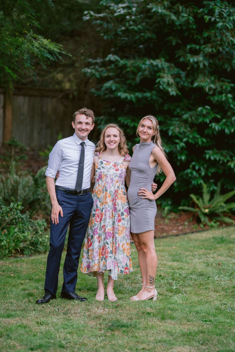 Three people pose together on a lawn with greenery in the background.